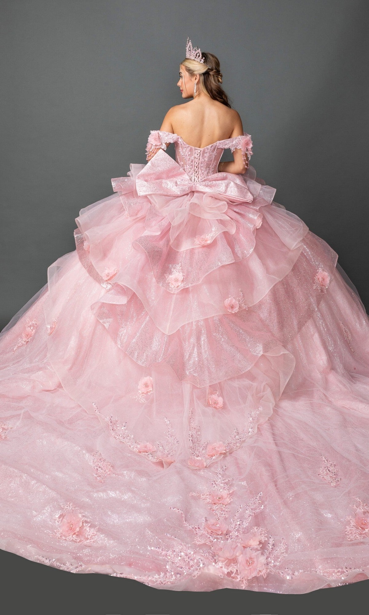 A woman in the Quinceanera Dress 5132 By Dancing Queen—a pink, off-the-shoulder ball gown with layered tulle, 3D flowers, a large back bow, and a tiara—stands facing away.